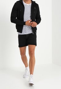 Sudadera negra con cremallera y capucha, llevada sobre una camiseta de rayas blanca y azul marino, combinada con pantalones cortos negros y zapatillas blancas.