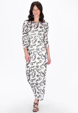 Kvinde iført en hvid maxikjole med lange ærmer med sorte blomstrede mønstre og sorte sandaletter med stropper, stående foran en ensfarvet baggrund.
