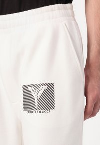 Biele športové nohavice majú na ľavom stehne čierne geometrické logo s textom. Tkanina sa zdá byť mäkká, má elastický pás a bočné vrecká.