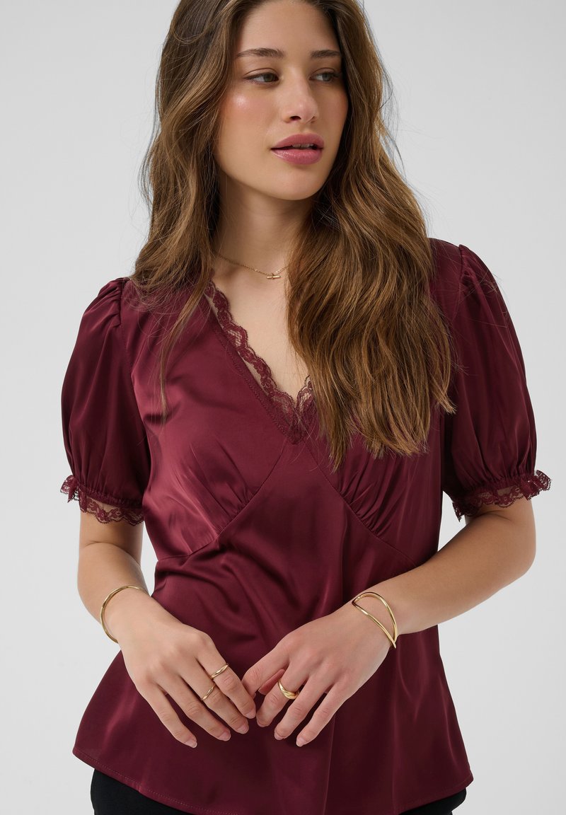 Jeune femme aux longs cheveux bruns portant un chemisier en satin bordeaux avec bordure en dentelle et des bijoux en or, regardant légèrement de côté.