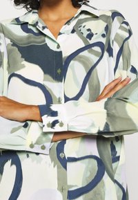 Una persona con capelli ricci indossa una camicia a bottoni con un motivo nei toni di verde e blu, con le braccia incrociate, mettendo in evidenza il design della camicia.