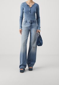 Ljusblå långärmad henley-top med tre knappar, kombinerad med vida, slitna ljusblå jeans. Modellen håller en blå handväska.