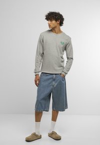Camisa gris de manga larga con logo verde, combinada con pantalones cortos de mezclilla hasta la rodilla. Zapatos beige sin cordones y calcetines blancos hasta el tobillo visibles.