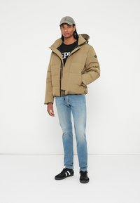 Kaki pufjakke med hætte og frontlynlås, parret med en sort grafisk t-shirt, lyseblå jeans og sorte sneakers.