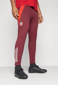 Bordeauxkleurige sportbroek met rode zijaccenten, met een Bayern München-logo. Drie witte strepen langs het onderbeen. Gladde, aansluitende stof.
