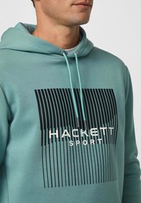 Sudadera con capucha en color verde azulado con diseño gráfico lineal negro. Cuenta con cordones y una textura suave, adecuada para uso casual.