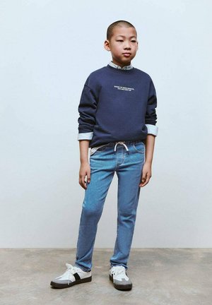 Hanorac bleumarin cu text alb, blugi albaștri deschiși și adidași albi cu accente gri. Design ajustat, texturi fine și stil casual.