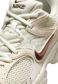 Primer plano de una zapatilla Nike beige con paneles de malla, superposiciones de ante, cordones blancos y un logotipo Nike Swoosh marrón.