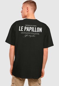Sort bomulds t-shirt med stor hvid tekst på ryggen: "LE PAPILLON" og et citat om kaosteori. Korte ærmer, afslappet pasform.