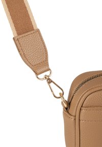 Ecke einer braunen Ledertasche mit goldenem Reißverschluss und Schnalle, die einen breiten gestreiften Stoffriemen befestigt.