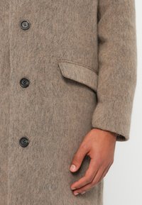 Närbild av en beige ullkappa med svarta knappar och en klaffficka, som visar bärarens underarm och hand.