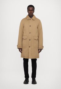 Tan coat with a collar, front buttons, and two front pockets. Features a straight cut and is paired with black pants and shoes.