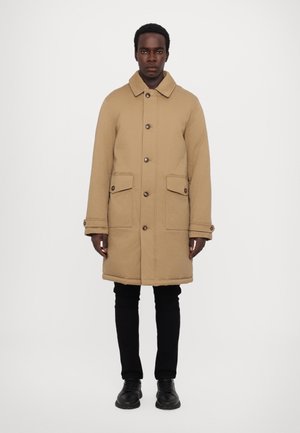 Tan coat with a collar, front buttons, and two front pockets. Features a straight cut and is paired with black pants and shoes.