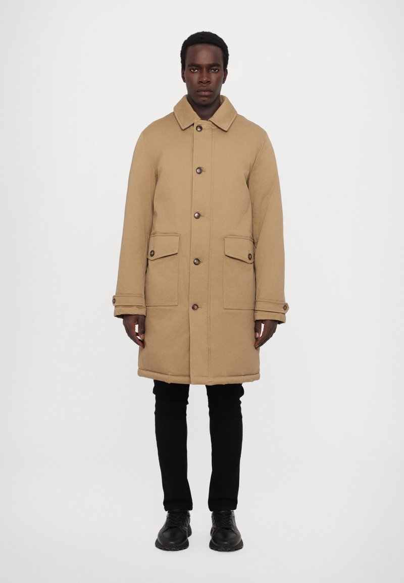 Tan coat with a collar, front buttons, and two front pockets. Features a straight cut and is paired with black pants and shoes.