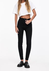 Jeans ajustados de tiro alto en negro, hechos de mezclilla, con una textura suave y diseño de cinco bolsillos, combinados con una camiseta corta blanca y zapatos negros.