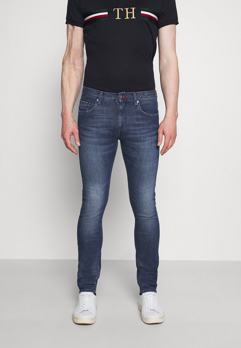 Homme portant un jean slim délavé bleu, des baskets blanches, et un t-shirt noir avec des rayures horizontales rouges, blanches et vertes ainsi que les initiales dorées "TH".