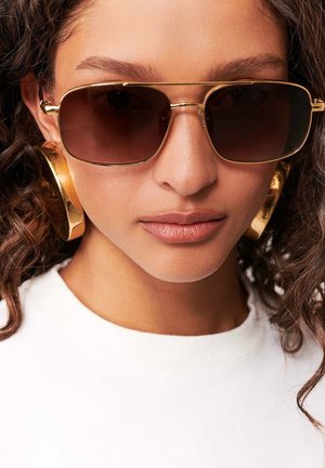 Kobieta z kręconymi włosami, nosząca brązowe okulary pilotki, duże złote kolczyki koła oraz biały top, zwrócona przodem.