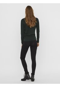 Femme aux longs cheveux bruns portant un pull texturé vert foncé, un jean skinny noir et des bottines noires à lacets, debout, de dos.