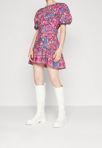 Vestido corto rosa con mangas abullonadas, patrón floral en azul y púrpura, cintura ajustada y un dobladillo con volantes. Combinado con botas blancas hasta la rodilla.