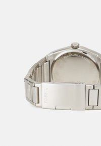 Montre en acier inoxydable avec un fini argenté poli. Elle présente un boîtier rond et un bracelet à maillons. Les inscriptions incluent "FOSSIL" et "TOUT EN ACIER INOXYDABLE."