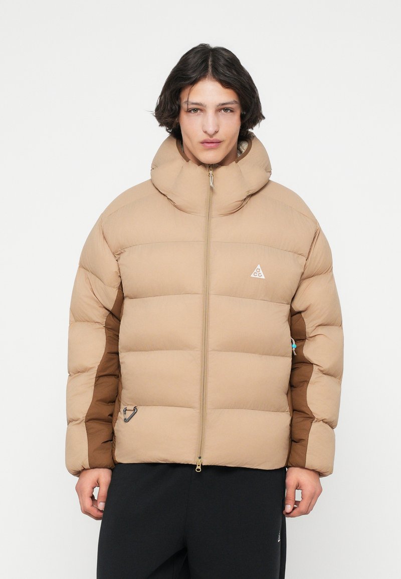 Chaqueta acolchada color beige con cuello alto, que presenta un cierre con cremallera en la parte delantera, mangas con detalles en color marrón y un logotipo. Textura suave con un ligero brillo.