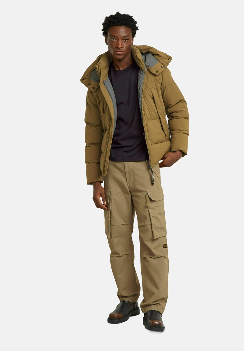 Khaki puchowy płaszcz z szarym wykończeniem, czarna koszula pod spodem i beżowe spodnie cargo. Model nosi czarne buty, ręce w kieszeniach.