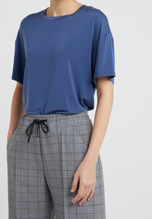 Persona que lleva una camisa azul suelta de manga corta metida dentro de unos pantalones anchos grises de cuadros con cordón negro, mano en el bolsillo.