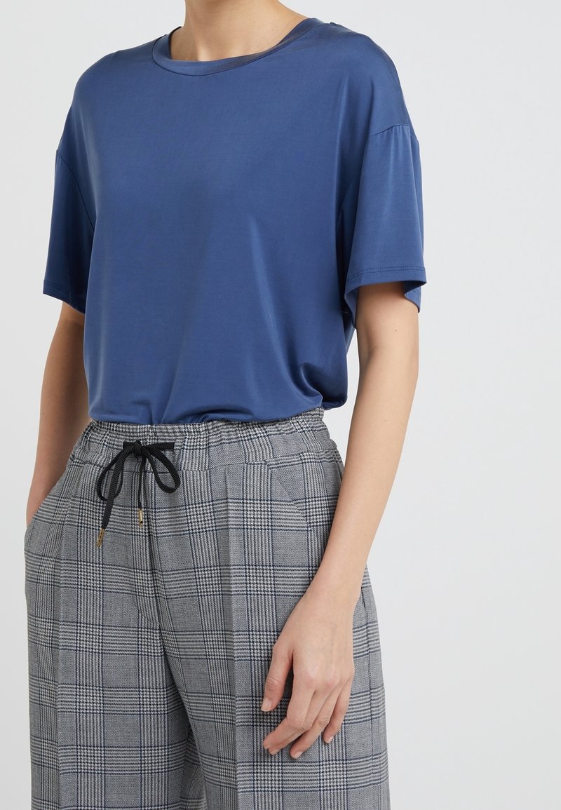 Personne portant une chemise bleue ample à manches courtes rentrée dans un pantalon gris à carreaux, jambes larges et cordon noir, main dans la poche.