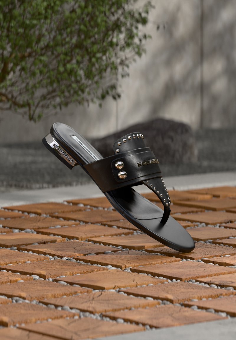 Μαύρο δερμάτινο σανδάλι με ασημένιες καρφίτσες και λογότυπο «Karl Lagerfeld», τοποθετημένο σε ξύλινα πλακάκια και χαλίκι σε εξωτερικό χώρο.