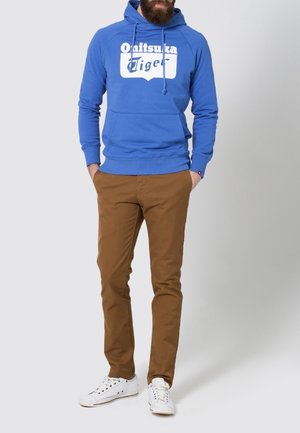 Blå bomullshuvtröja med vit "Onitsuka Tiger"-logga, kombinerad med bruna chinos och vita sneakers. Har en dragsnodd i huvan och ribbade muddarna.