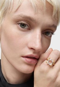 Anello d'oro con una pietra preziosa trasparente, caratterizzato da accenti sferici. Primo piano del volto di una persona con pelle chiara e capelli biondi corti.