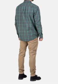 Chemise en flanelle à carreaux verts avec manches longues, ourlet incurvé et poignets à boutons, associée à un pantalon beige et des chaussures noires.