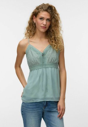 Femme aux cheveux bouclés portant un top sans manches vert clair orné de dentelle et un jean bleu, debout avec une main dans la poche sur fond blanc.