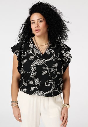 Vrouw met krullend zwart haar, gekleed in een zwarte blouse met wit bloemmotief, witte broek, gouden sieraden en armbanden, tegen een effen achtergrond.
