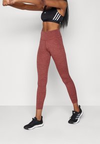 Leggings en bruyère rouge avec une taille haute, présentant une texture lisse et un design sans coutures. Associés à des chaussures de sport noires avec des accents blancs.