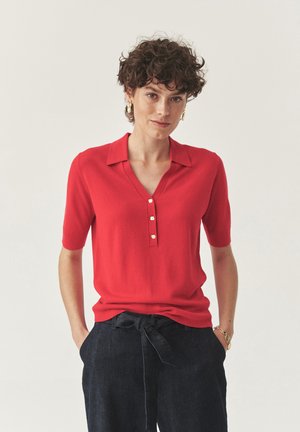 Polo shirt rosso a maniche corte con scollo a V e tre bottoni dorati. Il materiale appare morbido, con una vestibilità comoda e una trama liscia.