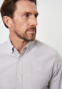 Camisa de botón gris claro de tela de algodón, con cuello, botones blancos y un pequeño logo bordado en el lado izquierdo del pecho.