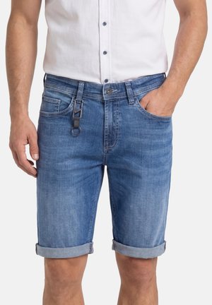 Mann trägt hochgekrempelte blaue Jeansshorts mit angebrachtem Schlüsselanhänger, kombiniert mit einem weißen Hemd, Hand in der Tasche.