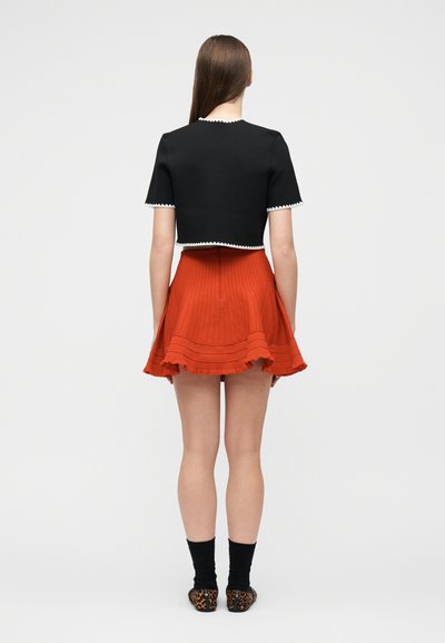 Γυναίκα με ίσια καστανά μαλλιά, που φοράει μαύρο κοντό τοπ με λευκή περικοπή και πορτοκαλί πλισέ φούστα, στέκεται με την πλάτη γυρισμένη, πάνω σε απλό φόντο.