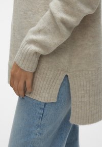 Pull en maille beige avec poignets et ourlet côtelés, fendu sur le côté, porté avec un jean en denim bleu clair.