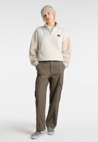 Pull col montant beige clair à fermeture éclair, agrémenté d'un logo, associé à un pantalon cargo vert olive avec poches latérales et des chaussures à enfiler à carreaux.