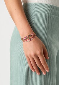 Multi-strand wrist bracelets in pink, gold, and silver featuring rectangular accents and letter beads spelling "LOVE"; worn on a textured light green fabric.