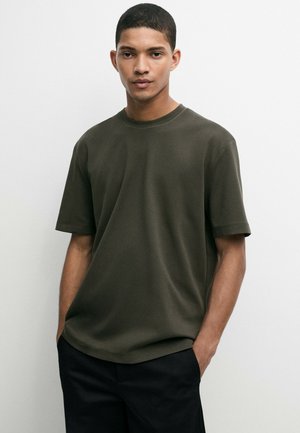 Ung mand med kort hår iført en mørkegrøn kortærmet t-shirt og sorte bukser, står foran en ensfarvet hvid baggrund.