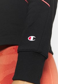 Svart sweatshirtärm med ribbad manschett, med en röd accentrand och en vit logotyp på armbågen. Bakgrunden visar en strukturerad röd textil.