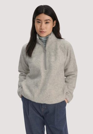 Vrouw met lang donker haar, gekleed in een lichtgrijze fleece trui met een kwart rits en donkerblauwe broek, handen in de zakken, neutrale achtergrond.