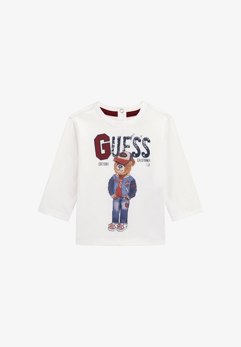 Camiseta blanca de manga larga con un gráfico de un oso vestido con chaqueta y gorra, con el logo "GUESS" y acentos rojos. Material de algodón suave.