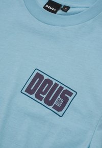 Light blue cotton t-shirt featuring a rectangular purple "DEUS" logo outlined in dark blue. Short sleeves and round neckline.
