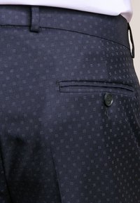 Pantaloni navy con un motivo testurizzato di piccoli puntini. Presentano una tasca piatta con bottone e una cintura liscia. Il tessuto sembra leggero.