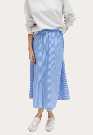 Kvinde iført en lysegrå sweatshirt, blå og hvide lodrette stribede midi-nederdel og hvide sneakers, står med den ene hånd i lommen.