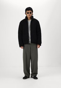 Veste en polaire noire et duveteuse avec col montant, associée à un pull gris à côtes, un pantalon olive vert décontracté et des chaussures noires à enfiler.
